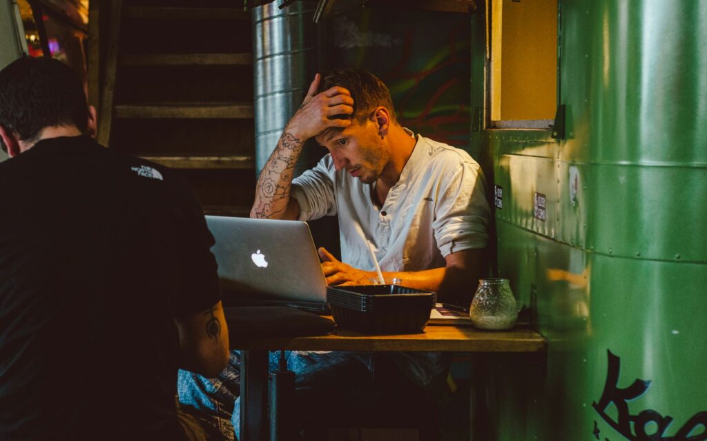 Frustrated tattooed business owner with hand on head looking stressed while working on laptop in industrial setting